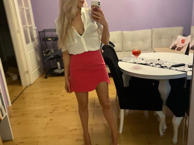 Lilith91-hot with blonde hair, white blouse showing her tits, red skirt, and heels, taking a mirror selfie in a room with a table, glass of red wine, and white couch.