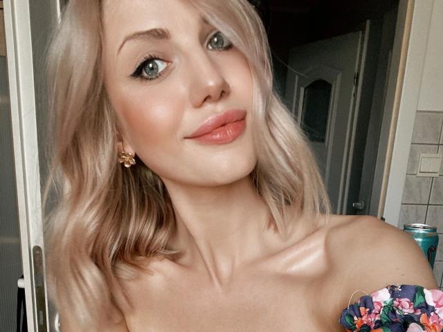 Lilith91-hot heeft blond haar, groene ogen en roze lippen met een subtiele glimlach. Ze draagt een bloemenprint top met een open schouder, waardoor haar borsten zichtbaar zijn. Ze heeft een schattige neus en kleine oorbellen..