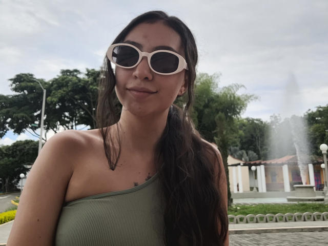 EmmaWells with long brown hair, wearing a green one-shoulder top, white sunglasses, and a slight smile, showing her small tits, standing outdoors with a fountain in the background.