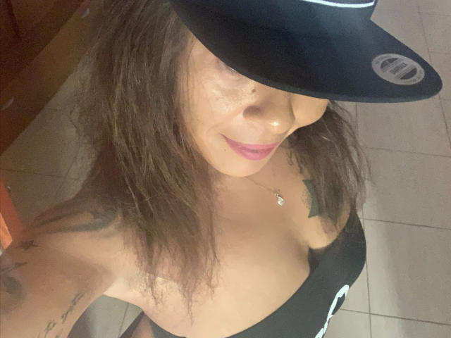Brownsugarheaven-hot with wavy brown hair, wearing a black hat and low-cut black top, showing her tits and tattoos, smiling with pink lipstick, tile floor below.