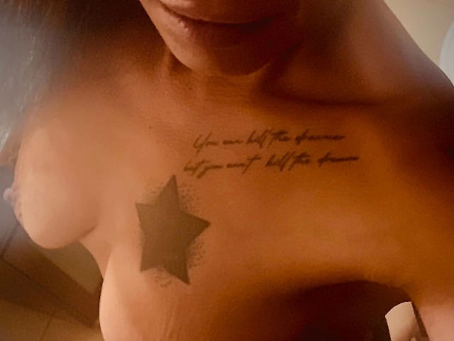 brownsugarheaven-hot with star tattoo on chest, cursive text above, medium boobs, tan skin, partial face visible, dim lighting, upper body shot, no clothes.