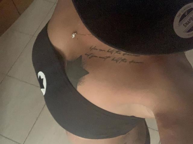 Brownsugarheaven-hot's lower torso, wearing black underwear with a white moon logo, showing a tattoo above their tits and a navel piercing, standing on tiled floor.