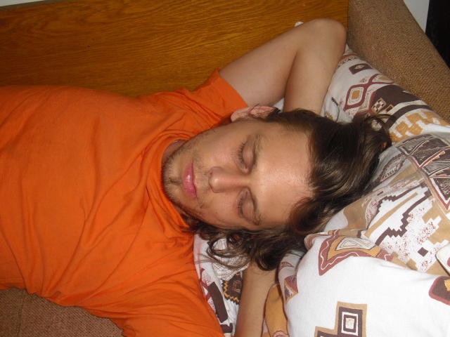 OndraSekCZech is sleeping with eyes closed, wearing an orange shirt, and has brown hair. His arm is resting on a patterned pillow. He's on a brown couch.