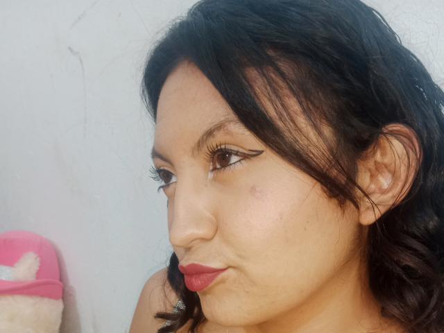 SalomeHottieX has medium brown skin, dark wavy hair, and is pouting with red lipstick, wearing black eyeliner. Her nose has a small blemish on the right side.
