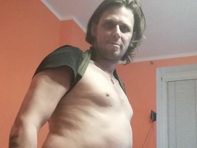 OndraSekCZech with shoulder-length brown hair, shirtless, showing a toned, pale chest with small nipples, and a subtle smile, standing in a room with orange walls.