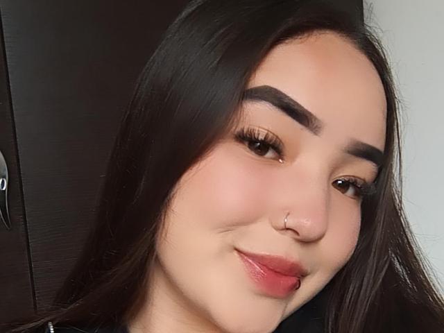 LilaRosa69 has dark brown hair, thick eyebrows, long eyelashes, a small nose ring, and a slight smile with pink lips. Her skin is light, and she's looking to the side.