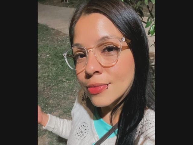 LindaLuna with tan skin, black hair, and clear glasses, sticking her tongue out, wearing a white cardigan over a teal shirt, and a black strap.