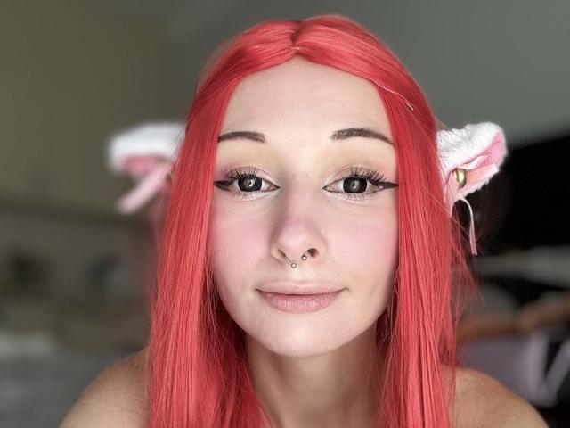 HannaCool, avec ses cheveux roux, ses oreilles de chat et son anneau de nez, souriant à la caméra, mettant en valeur son eye-liner et ses boucles d'oreilles, avec une expression subtile et mignonne..