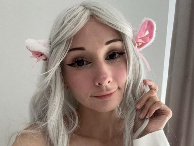 HannaCool has white hair, pink cat ears, and a nose ring. She's smiling with a hand in her hair, showing off her eyeliner and pink lips.