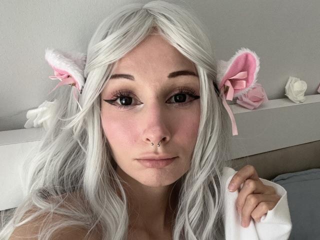 HannaCool with white hair, pink cat ears, and a nose ring, holding a white pillow, showing off her tits and ass in a selfie. Her makeup is cute and she's sexy.