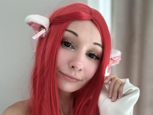 HannaCool with red hair, pink cat ears, and nose ring, smiling at camera, holding white cloth, cute face, pink lipstick, eyeliner, and small nose piercing.