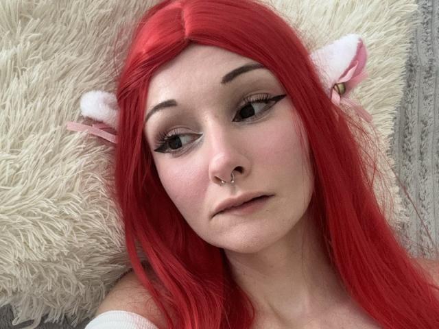 HannaCool with red hair, white furry ears, nose piercing, and winged eyeliner, looking at camera with a slight pout, lying on fluffy white pillow.