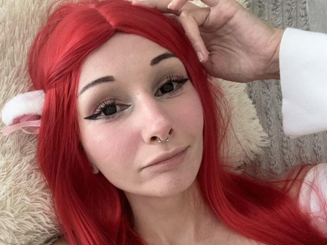 HannaCool with red hair, black eyeliner, nose ring, and a hand in her hair, looking at the camera with a slight smile, lying on a fluffy beige surface.