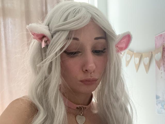 HannaCool with white, wavy, cat-ear wig, pink collar, nose ring, and heart necklace, looking down with closed eyes, pink blush, and subtle makeup, in soft lighting.