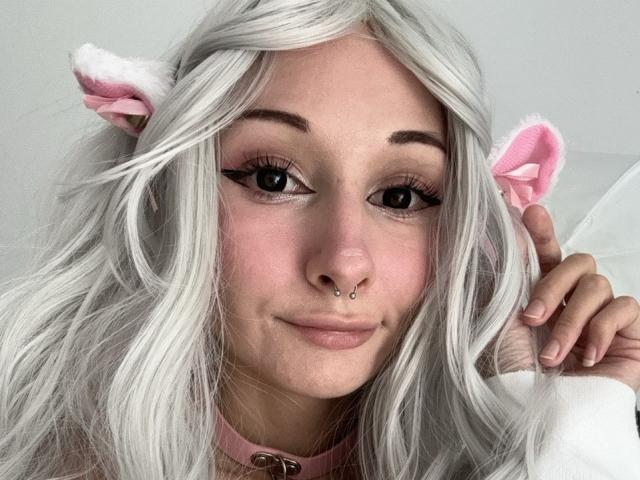 HannaCool with white hair, pink cat ears, nose piercing, and pink choker, smiling, showing off her tits, cute, and fluffy. Soft pink makeup, long lashes, and a gentle smile.