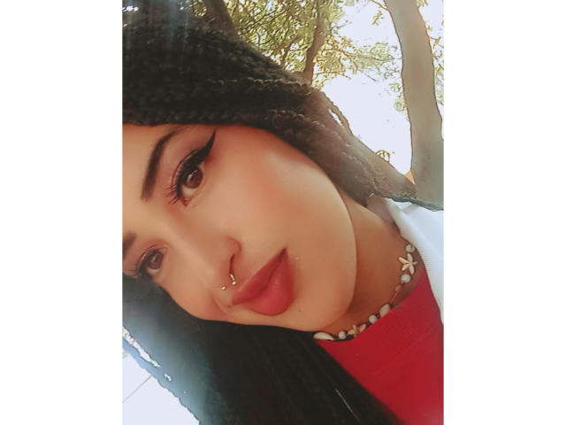 MichellDiaz has dark braids, a nose ring, and thick eyeliner, wearing a red shirt and white jacket, with a pearl necklace, and a sexy, confident smile.