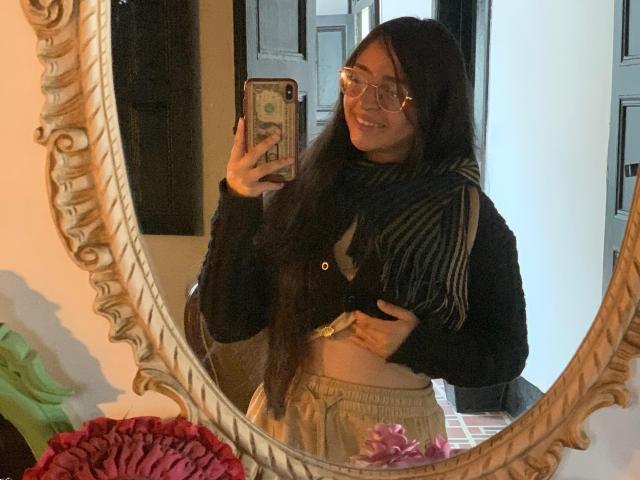 EvaMcAllister with long black hair, glasses, black sweater, striped scarf, beige pants, holding phone in mirror selfie, smiling, showing belly button, mirror with ornate frame.