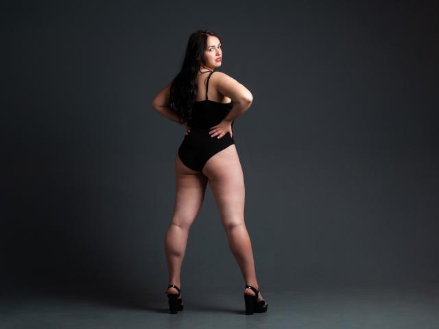 IsabelBlackberry with long black hair, wearing a black one-piece, standing in high heels, showing her ass, looking back with hands on hips, in a dark studio.
