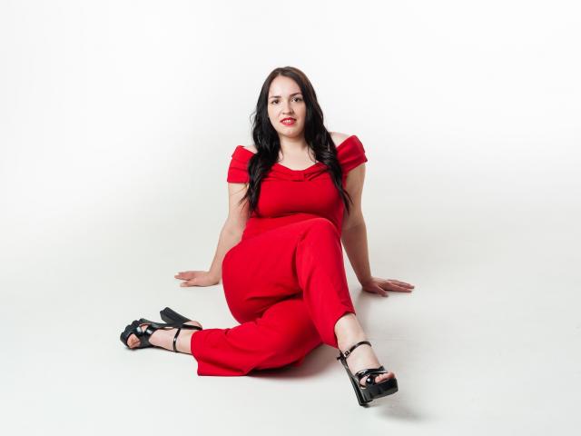 IsabelBlackberry, con cabello negro largo, vestido rojo y tacones altos negros, sentada en el suelo blanco, con las piernas separadas y los brazos detrás, labios rojos, mirando a la cámara, con una expresión segura..