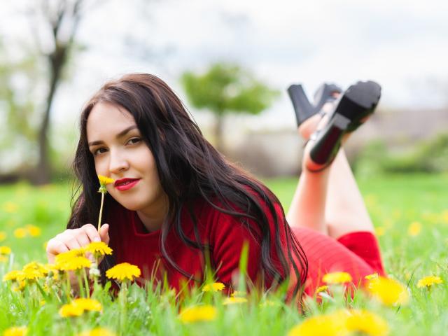 IsabelBlackberry with long dark hair, red dress, and black heels, lying in a green field, holding a yellow daisy, smiling, her ass up, legs crossed, in a sunny setting.