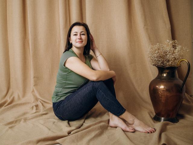 MarieSho, assise sur un tissu beige, pieds nus, chemise verte, jean foncé, souriante, cheveux bruns, main sur la tête, vase en cuivre avec des fleurs séchées à sa droite..