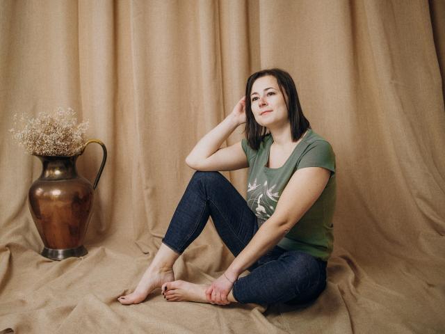 MarieSho with shoulder-length brown hair, green t-shirt, and dark jeans, sitting barefoot on beige fabric, hand on head, small smile, brown vase with dried flowers to the left.