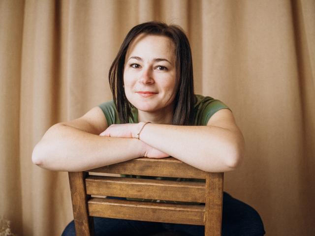 MarieSho with medium tits, arms crossed on wooden chair, green shirt, brown hair, slight smile, beige curtain background, bracelet on wrist, sitting, casual, relaxed.