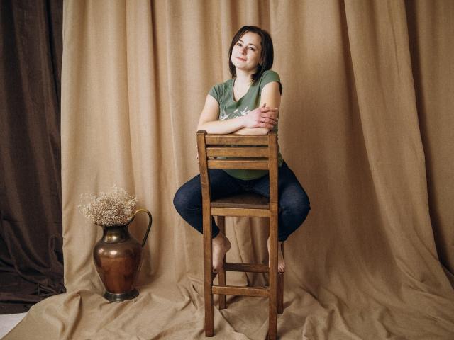 MarieSho sitting on a wooden chair, arms crossed, green shirt, blue jeans, bare feet, brown hair, neutral expression, brown vase with dried flowers to the left.