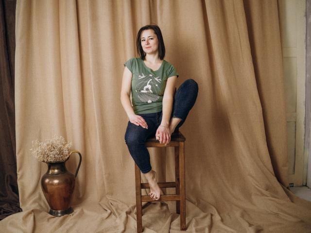 MarieSho, met één been op een houten kruk, gekleed in een groene blouse en blauwe jeans, haar blote voet op de kruk, bruin haar en een rustige uitdrukking, naast een vaas..