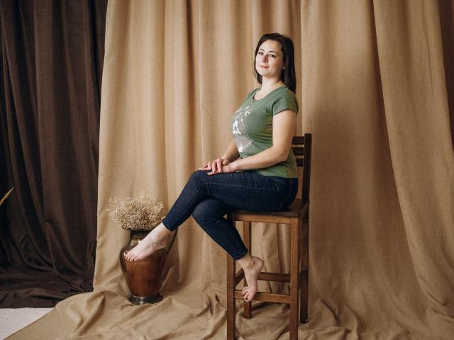 MarieSho with medium tits, green tee, dark jeans, bare feet on wooden chair, brown curtain backdrop, bronze vase with dry grass, sitting, arms on knees, soft smile.