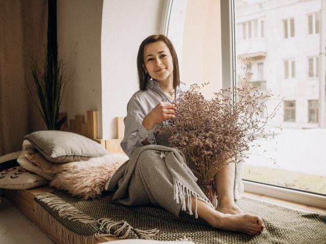 MarieSho with small tits, sitting on a window ledge, holding dried flowers, wearing a gray shirt and checkered skirt, smiling, barefoot, on a green rug with pillows.