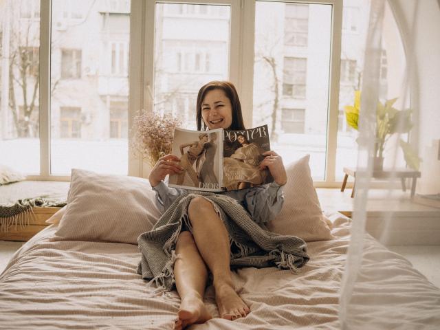 MarieSho with bare feet, sitting on a bed, wearing a blue dress, holding a magazine, smiling, light brown hair, white pillows, gray blanket, window in background.