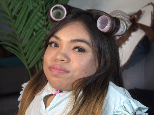 MichelleBrooks has brown hair in pink curlers, light skin with a small zit on her cheek, and wears a white shirt with a pink choker, smiling slightly.