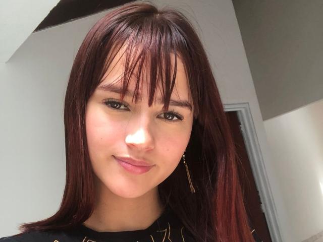 MelodyMansson has straight, dark red hair with bangs, brown eyes, and a slight smile. She's wearing a black top with gold details and gold earrings.