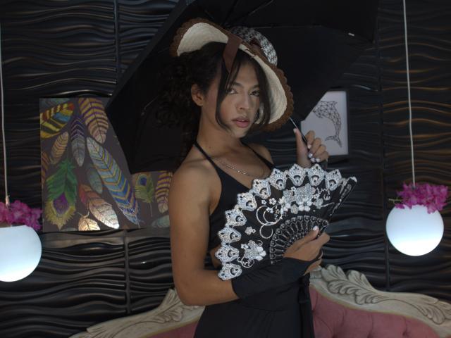 BriyitTs with dark curly hair, wearing black dress, holding black umbrella, white floral fan, black gloves, and white hat, standing in dimly lit room, looking at camera.