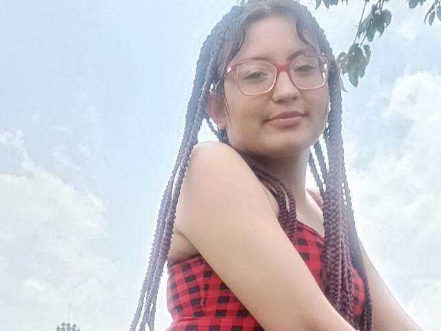 SalomeHottieX has long braided hair, red glasses, and a red plaid top, showing off her small tits and cute smile against a cloudy sky.