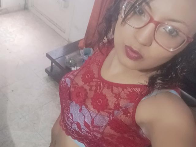 SalomeHottieX with red lace top, showing her tits, wearing glasses, red lipstick, and curly dark hair, in a selfie, looking at the camera.