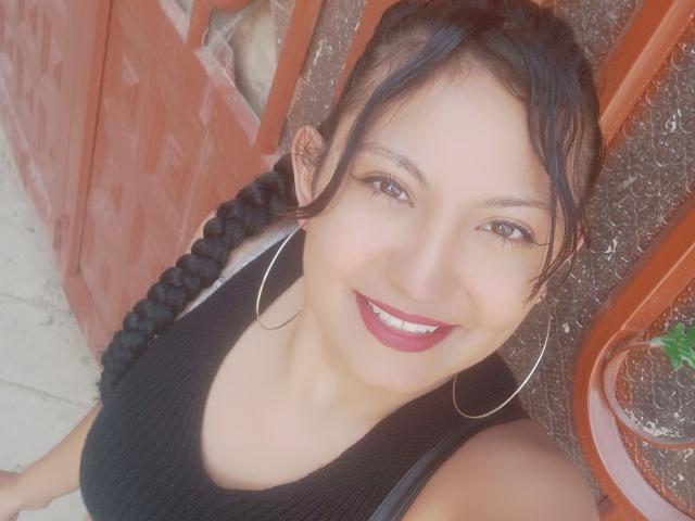 SalomeHottieX with dark braids, hoop earrings, and red lipstick, smiling at the camera, wearing a black ribbed tank top, showing her tits slightly.