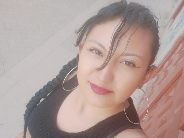 SalomeHottieX with dark hair in a braid, wearing a black top, hoop earrings, and purple lipstick, smiling at the camera. She has cute tits and a cute ass.