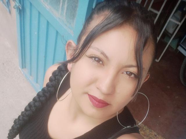 SalomeHottieX has light brown skin, dark braided hair, red lipstick, and large silver hoop earrings. She's wearing a black tank top and looking up at the camera.