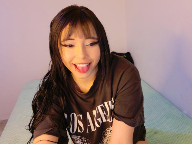 Arieyaara, with long black hair and bangs, sticks out her tongue, wearing a brown Los Angeles t-shirt, on a green bed, smiling at the camera.