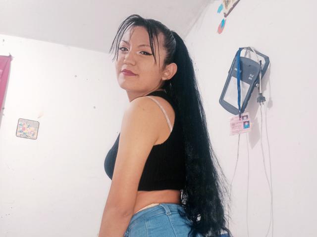 SalomeHottieX with long black hair in a ponytail, wearing a black crop top and blue jeans, showing her back and smiling over her shoulder. Her tits are small.