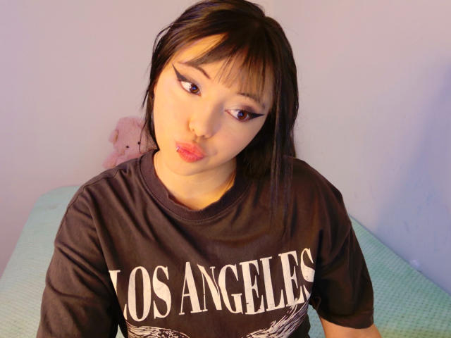 Arieyaara, con capelli neri, eyeliner rosa e piercing al naso, labbra tumefatte, indossa una maglietta marrone con la scritta "Los Angeles", seduta su un letto verde, con un peluche rosa sullo sfondo..