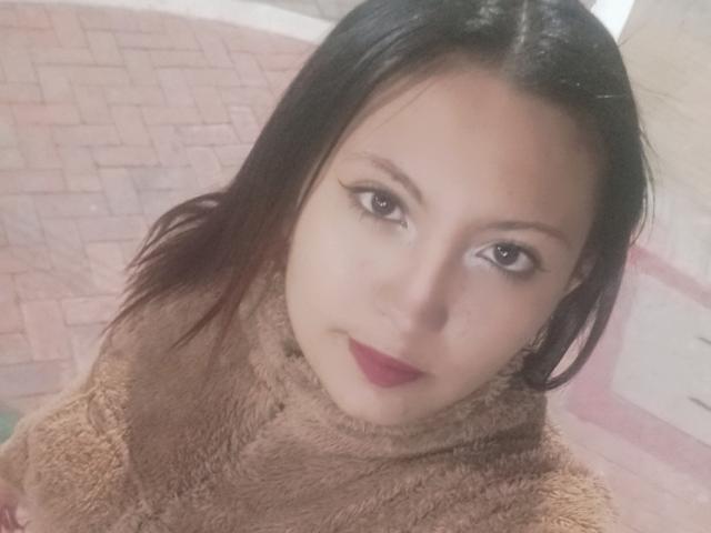 SalomeHottieX with brown eyes, black hair, and red lipstick, wearing a fuzzy brown turtleneck, looking up with a slight smile, her cute face and small nose visible.