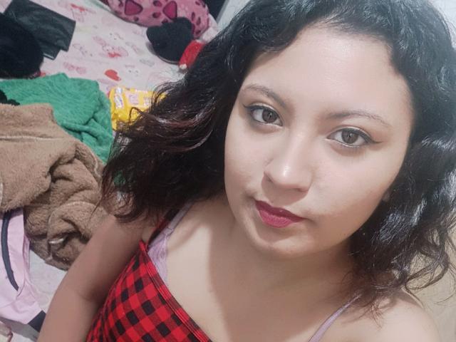 SalomeHottieX has medium brown skin, wavy black hair, and red lipstick. She's wearing a red and black checkered top, showing her small tits. She's looking at the camera.
