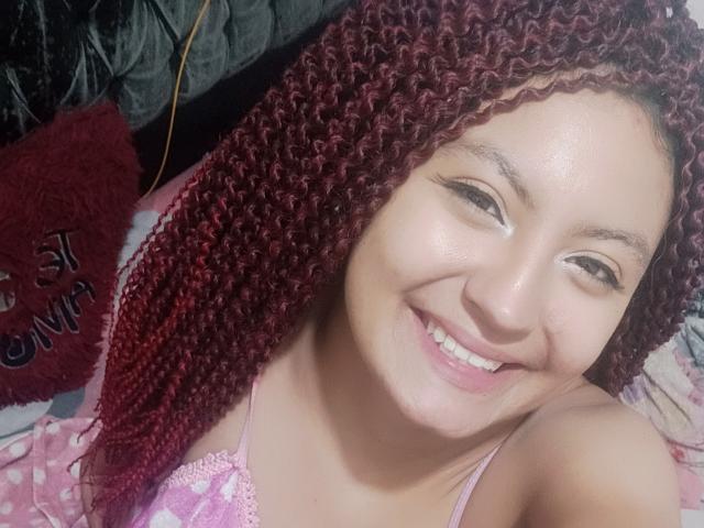 SalomeHottieX with red curly hair, smiling, showing teeth, wearing pink lace top, pink polka dot background, white skin, brown eyes, looking at camera, cute, attractive.