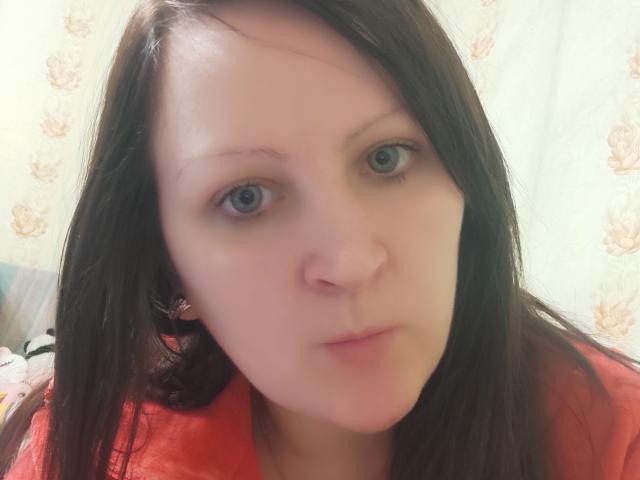 MillaOne has pale skin, blue eyes, and long dark brown hair. She's wearing a red shirt, looking slightly off-camera with a neutral expression. Her nose is small.
