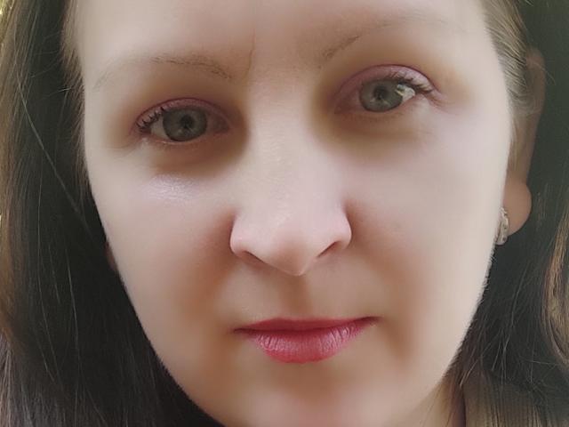 MillaOne's face, showing her pale skin, green eyes, pinkish-brown eyelids, thin eyebrows, small nose, pink lips, and straight brown hair, with a slight smile.