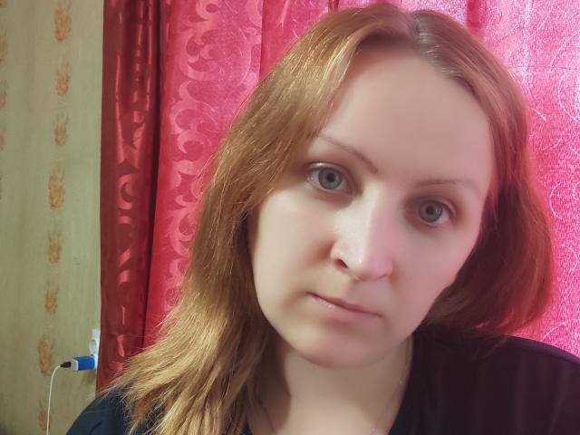 MillaOne has pale skin, red hair, and green eyes. She's looking directly at the camera with a neutral expression, wearing a black top. Her nose is slightly upturned.