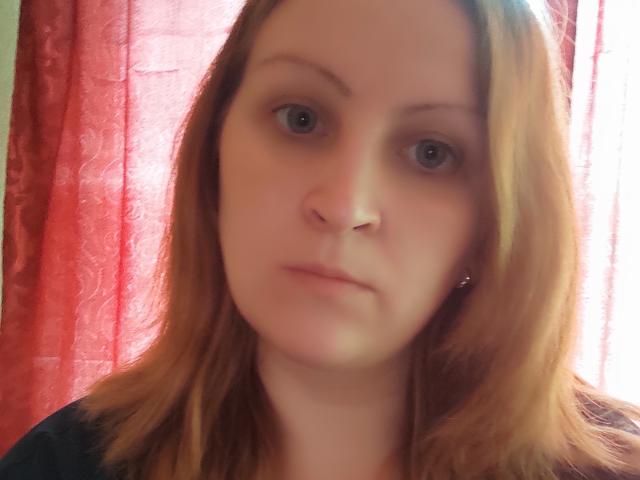 MillaOne has pale skin, blue eyes, and brown shoulder-length hair, with a slightly blurred face and a neutral expression, wearing a dark shirt, standing in front of red curtains.
